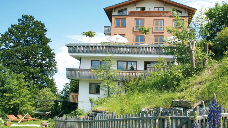 Ein mehrst&ouml;ckiges Hotel mit Holzbalkonen, umgeben von gr&uuml;ner Natur und bl&uuml;henden Blumen, unter einem blauen Himmel.