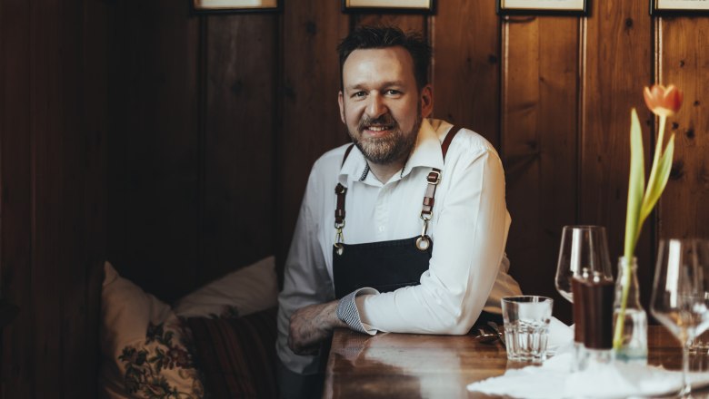 Ein Mann in einem Restaurant sitzt an einem Holztisch, trägt eine weiße Bluse und eine Schürze. Im Vordergrund steht eine Tulpe in einer Vase.