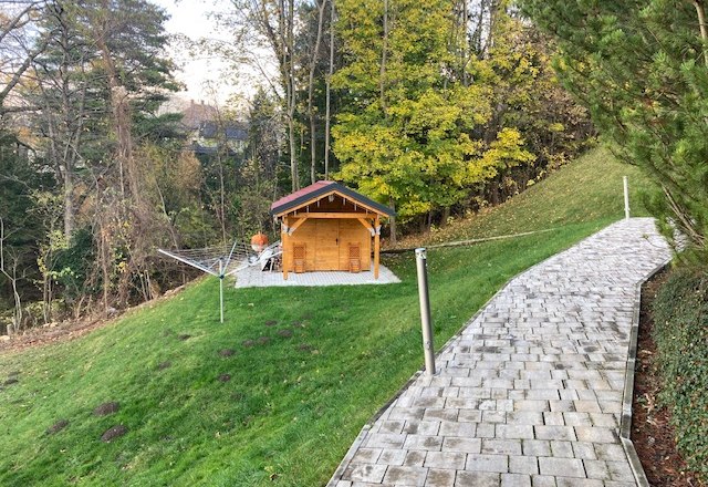 Ein Garten mit einem kleinen Holzhaus, umgeben von B&auml;umen und einem gepflasterten Weg.