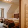 Gem&uuml;tliches Hotelzimmer mit Doppelbett aus hellem Holz, Fenster mit Vorh&auml;ngen und einem Sessel. Im Vordergrund sieht man ins Badezimmer. Das Zimmer hat fensterseitig eine Dachschr&auml;ge.