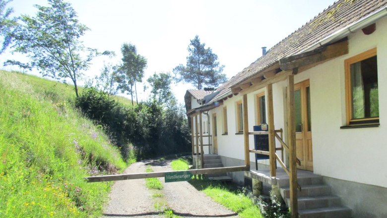 Weißes Gebäude mit einem Schranken mit dem Schild "Mostheuriger"  neben einem Schotterweg und einer grünen Wiese mit Bäumen.