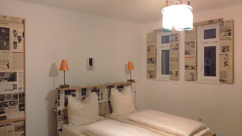 Hotelzimmer mit Doppelbett, Zeitungsdekoration an W&auml;nden und Fenstern, zwei orangefarbene Lampen.
