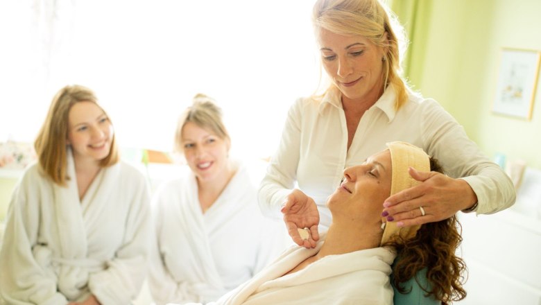Eine Kosmetikerin behandelt das Gesicht einer Frau. Zwei andere Frauen im Bademanteln sehen zu.