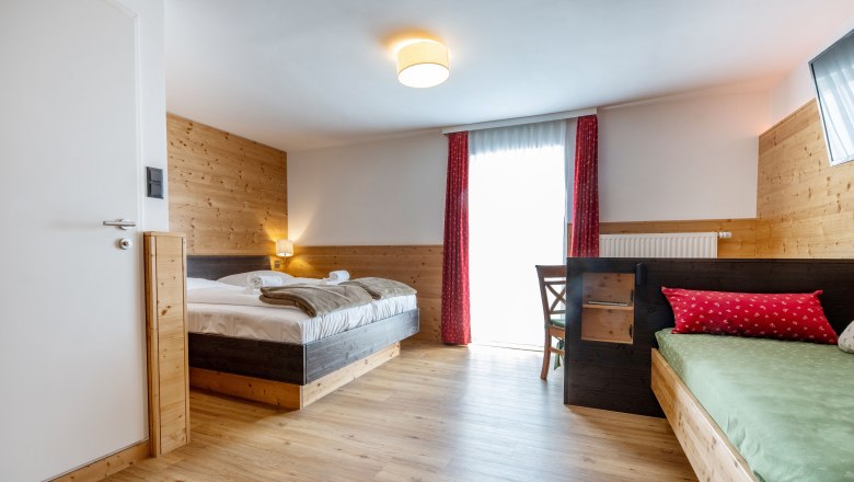 Gem&uuml;tliches Hotelzimmer mit Holzw&auml;nden, Doppelbett, Einzelbett und rotem Vorhang.