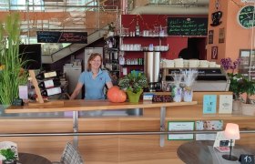 Eine Frau steht hinter der Theke eines Caf&eacute;s mit Pflanzen, Weinflaschen und einer Kaffeemaschine.