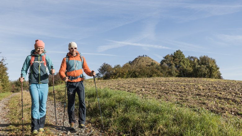 Zwei Wanderer auf einem Pfad in einer ländlichen Landschaft mit Hügel im Hintergrund.