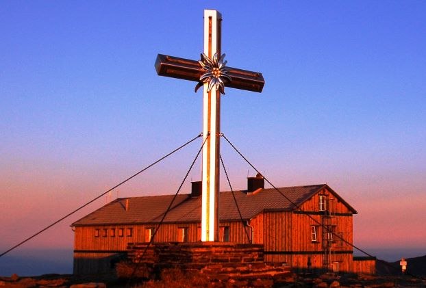 Großes Gipfelkreuz vor dem Alois-Günther-Haus bei Sonnenuntergang.