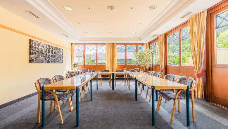 Ein heller Seminarraum mit Tischen in U-Form, St&uuml;hlen und gro&szlig;en Fenstern mit Vorh&auml;ngen.