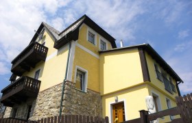 Gelbes Haus mit Holzdetails und Steinmauer, blauer Himmel im Hintergrund.