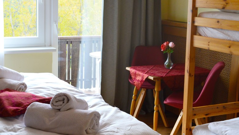 Ein gem&uuml;tliches Zimmer mit einem Bett, einem Tisch mit roten St&uuml;hlen und einem Blumenstrau&szlig;. Ein Fenster zeigt einen Balkon und herbstliche B&auml;ume.
