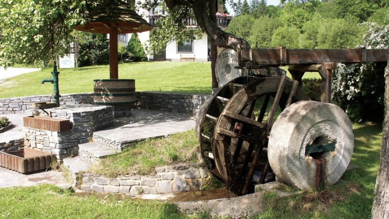 Altes Mühlrad mit Mühlstein, daneben Stufen und ein Brunnen, umgeben von Wiese und Bäumen.