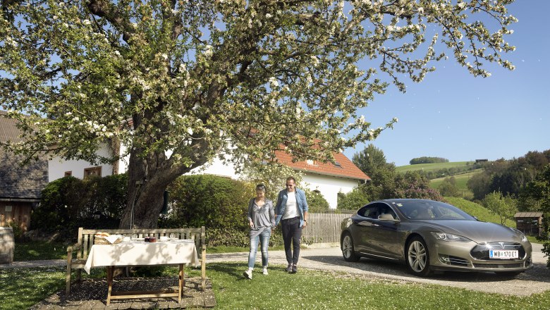Ein Paar geht neben einem geparkten Elektroauto unter einem blühenden Baum in einem ländlichen Garten.