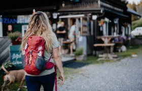 Frau mit rotem Rucksack und Hunde geht auf eine Bergh&uuml;tte zu.