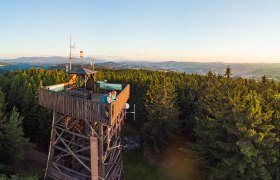Aussichtsturm aus Holz im Wald mit zwei Personen, die den Sonnenuntergang betrachten, im Hintergrund h&uuml;geliges Panorama,