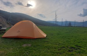 Ein orangefarbenes Zelt steht auf einer gr&uuml;nen Wiese, umgeben von Bergen und einem Zaun, bei bew&ouml;lktem Himmel und Sonnenuntergang.