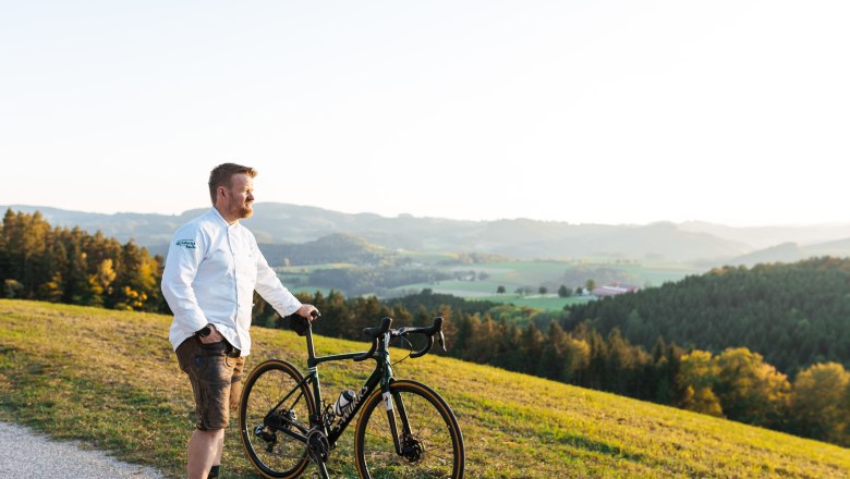 Ein Mann in wei&szlig;em Hemd steht mit einem Fahrrad auf einem H&uuml;gel mit Blick auf eine gr&uuml;ne Landschaft.
