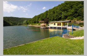 Ein See mit einem gelben Gebäude am Ufer, umgeben von grünen Hügeln und Bäumen.