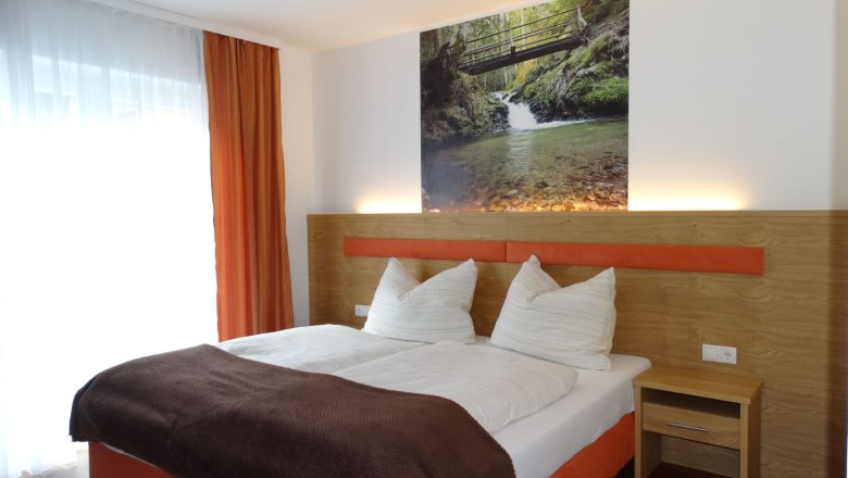 Hotelzimmer mit Doppelbett, orangen Vorhängen und Naturbild an der Wand.