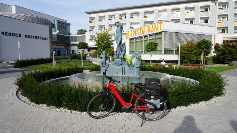 Brunnen mit einer Statue vor einem Geb&auml;ude mit der Aufschrift "Kurrestaurant", davor steht ein rotes Fahrrad..