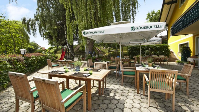 Ein sonniger Gastgarten mit Holzm&ouml;beln und Sonnenschirmen, umgeben von gr&uuml;ner Vegetation und einem gelben Geb&auml;ude.