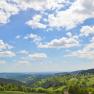 Panoramablick auf eine h&uuml;gelige Landschaft mit W&auml;ldern und Wiesen unter einem blauen Himmel mit wei&szlig;en Wolken.
