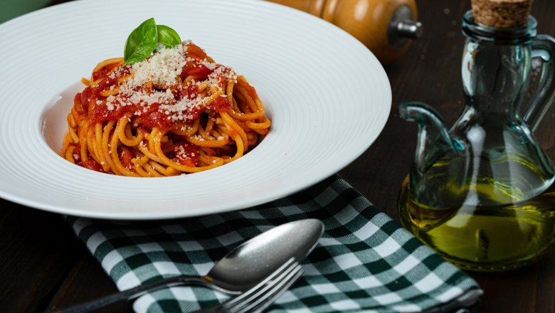 Ein Teller Spaghetti mit Tomatensauce, Basilikum und Parmesan, daneben eine &Ouml;lflasche und Besteck auf einem karierten Tuch.