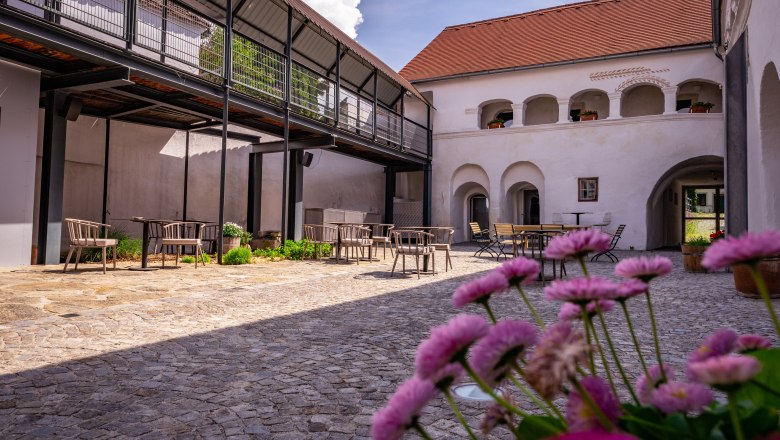 Ein Innenhof mit Kopfsteinpflaster, Tischen und Stühlen, umgeben von Gebäuden mit Arkaden und einem modernen Balkon.