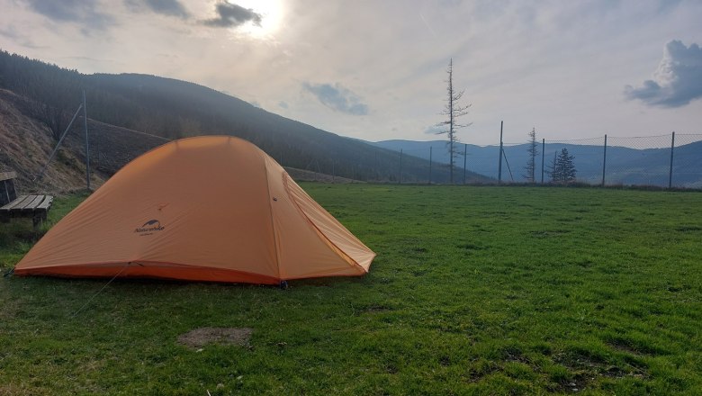 Ein orangefarbenes Zelt steht auf einer gr&uuml;nen Wiese, umgeben von Bergen und einem Zaun, bei bew&ouml;lktem Himmel und Sonnenuntergang.