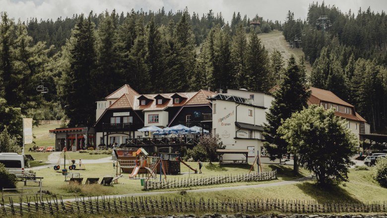 Ein Gasthaus in einer bergigen Waldlandschaft mit Spielplatz und Seilbahn im Hintergrund.