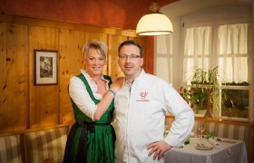 Ein Mann und eine Frau stehen in einem gemütlichen Restaurant. Die Frau trägt ein Dirndl, der Mann eine Kochjacke mit der Aufschrift 'Zum Forsthaus'.