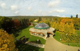 Luftaufnahme eines runden Gebäudes mit Glaskuppel, umgeben von herbstlichen Bäumen und einem Garten.