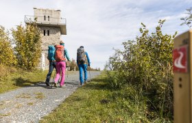 Drei Wanderer mit Rucksäcken gehen auf einem Weg zu einem Steinturm.