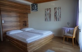 Ein Schlafzimmer mit einem Doppelbett, Holzwandverkleidung, zwei abstrakten Gemälden und einem kleinen Tisch mit Stuhl.