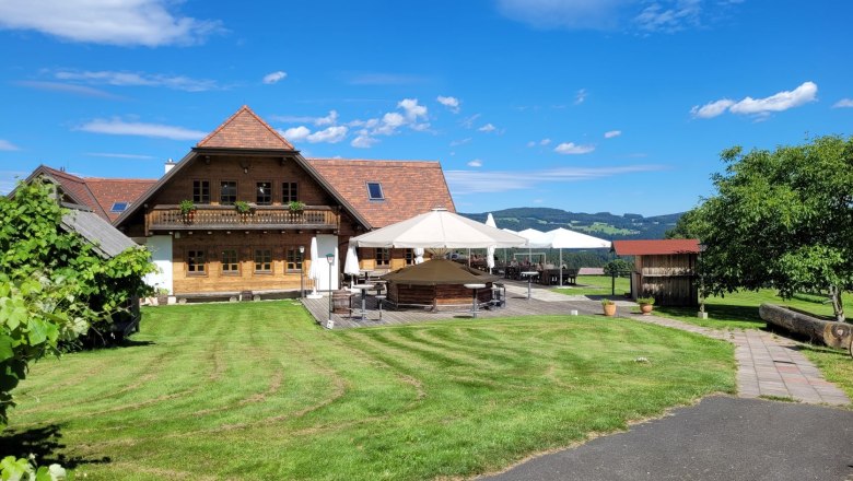 Ein traditionelles Holzhaus mit Terrasse und Sonnenschirmen in einer grünen Landschaft unter blauem Himmel.