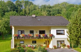 Zweist&ouml;ckiges gelbes Haus mit Balkon und Blumen, umgeben von B&auml;umen und Garten.