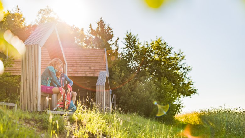 Zwei Personen sitzen in einer h&ouml;lzernen Struktur in einer sonnigen Landschaft.