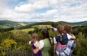 Das Ehepaar Berger, die beiden T&ouml;chter am Arm, blickt in die Landschaft mit H&uuml;geln und W&auml;ldern.