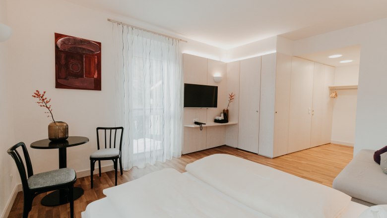 Modernes, helles Hotelzimmer mit Doppelbett, TV, Tisch und St&uuml;hlen, Schrank und dekorativem Bild an der Wand.