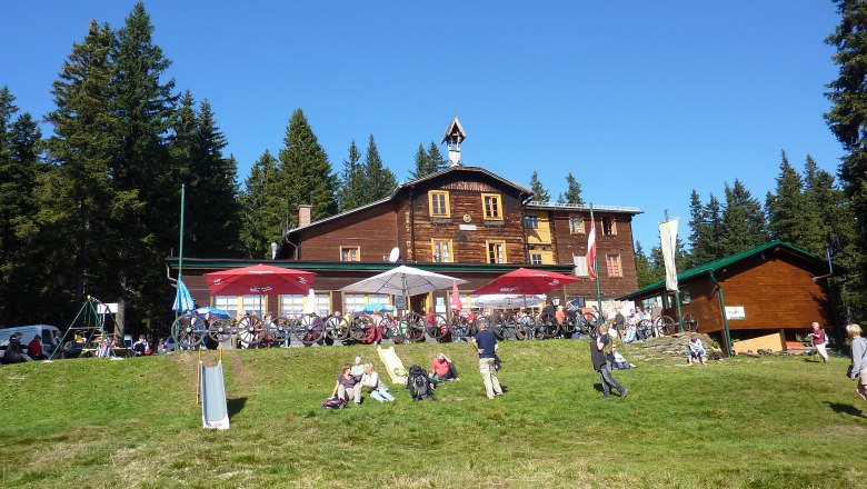 Haller-ház, © Hallerhaus, Foto Heinz Bayer Faház a hegyekben, előtte emberek és kerékpárok, fákkal körülvéve.