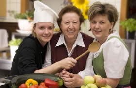 Drei Frauen in einer Küche, eine trägt eine Kochmütze, Gemüse und Obst im Vordergrund.