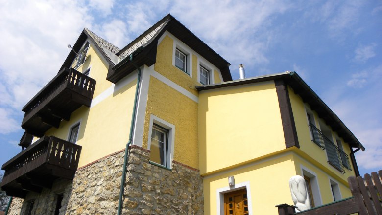 Hausansicht, © Thomas Baldauf Gelbes Haus mit Holzdetails und Steinmauer, blauer Himmel im Hintergrund.