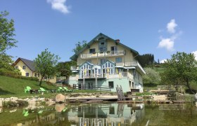 Ein großes, gelbes Haus mit blauen Akzenten spiegelt sich in einem Teich. Im Vordergrund sind grüne Liegestühle und Bäume zu sehen.