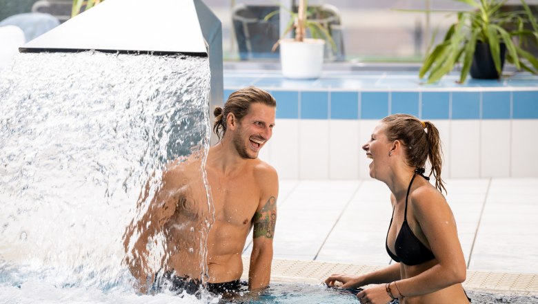Ein Mann und eine Frau stehen lachend in einem Pool unter einem Wasserfall.