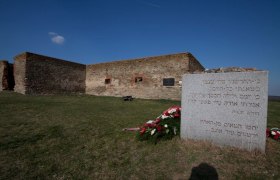 Gedenkstein mit hebr&auml;ischer Inschrift vor den Resten eines kreuzf&ouml;rmigen Steingeb&auml;ude.