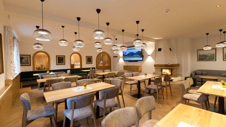 Ein gem&uuml;tliches Kaminzimmer mit Holztischen, St&uuml;hlen und modernen Lampen. Ein Kamin brennt im Hintergrund.