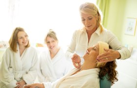 Eine Kosmetikerin behandelt das Gesicht einer Frau. Zwei andere Frauen im Bademanteln sehen zu.