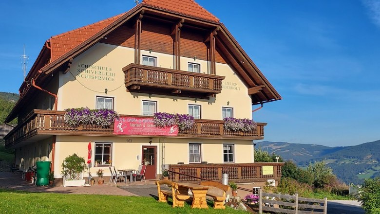 Pension Koderholt, © Koderholt Ein traditionelles Gasthaus mit Holzbalkonen und Blumen, umgeben von grünen Hügeln und blauem Himmel.
