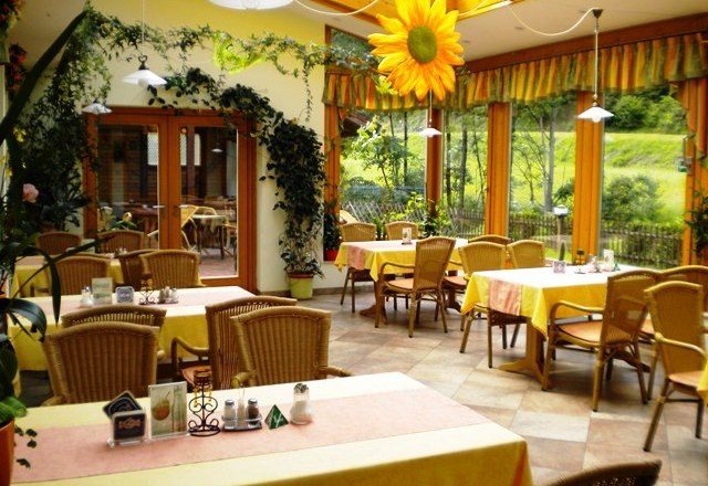 Ein gem&uuml;tlicher Wintergarten mit gelben Tischdecken, Rattanm&ouml;beln und Pflanzen. Gro&szlig;e Fenster bieten Blick ins Gr&uuml;ne. Eine gro&szlig;e Sonnenblume h&auml;ngt von der Decke.