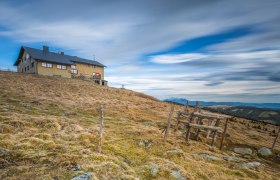 Das Wetterkoglerhaus auf einem H&uuml;gel mit bew&ouml;lktem Himmel im Hintergrund.