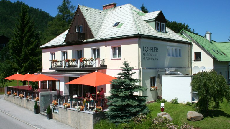 Ein zweist&ouml;ckiges Geb&auml;ude mit der Aufschrift 'L&ouml;ffler Pension-Caf&eacute;-Restaurant', umgeben von B&auml;umen und Blumen, mit orangenen Sonnenschirmen auf der Terrasse.